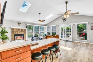A spacious kitchen with a large island, a fireplace, and a dining area with bar stools. There are ceiling fans and large windows providing natural light.