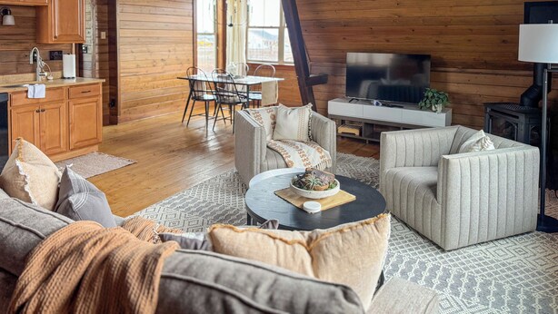 A cozy cabin interior with wooden walls, a dining area, a living room with sofas and a coffee table, and a fireplace. - Creston Centennial #1-King bed, rustic barn loft w/stairs (Kalispell)
