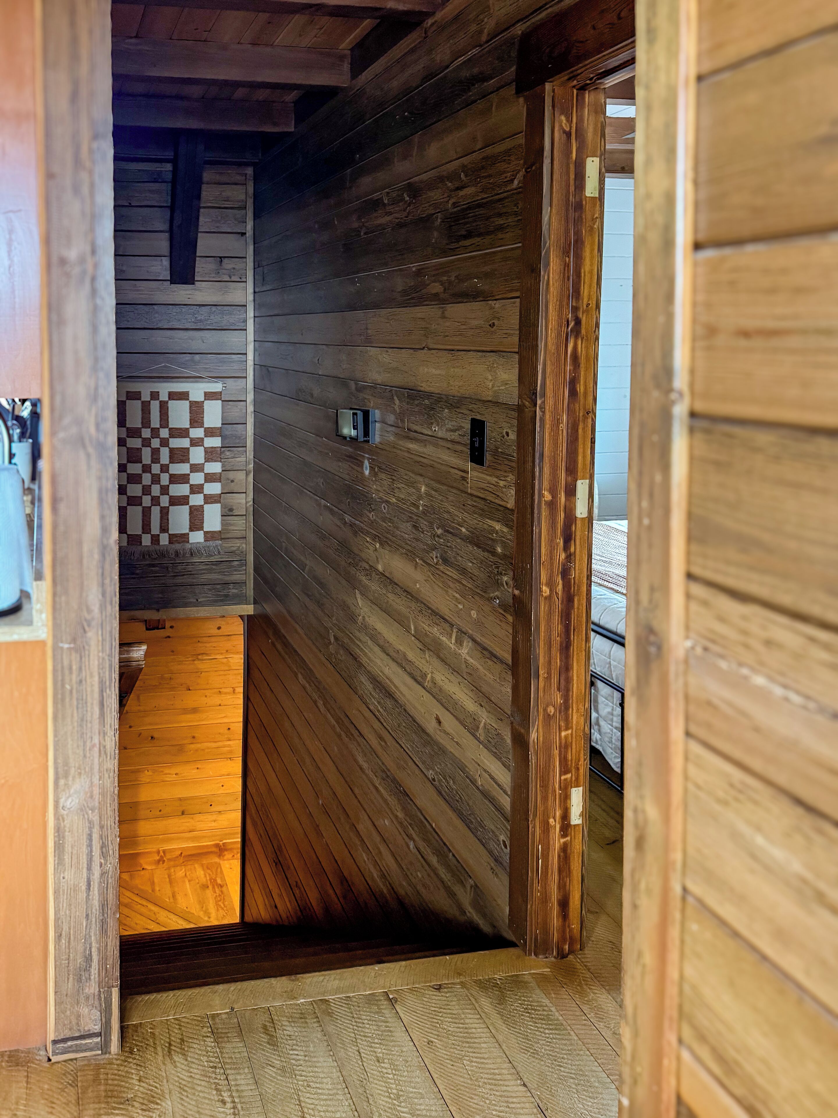 A wooden interior hallway with a checkered wall hanging, a door with a light switch, and a bed with a white comforter.