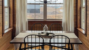 A dining room with a wooden table, four chairs, a chandelier, and a view of a field through large windows. - Creston Centennial #1-King bed, rustic barn loft w/stairs (Kalispell)