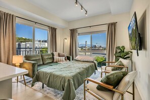 A modern bedroom with a large bed, a desk, and a chair. There are two windows with curtains, a TV on the wall, and a plant.