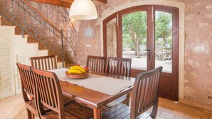 Un comedor con una mesa de madera, sillas y un frutero. Se ve una escalera con barandilla y una puerta que da al exterior.