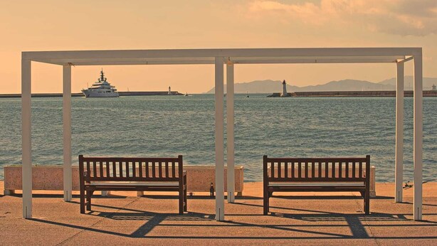 A waterfront bench area with a view of the sea, a boat, and a lighthouse in the distance.