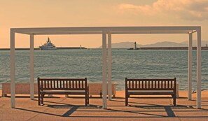 A waterfront bench area with a view of the sea, a boat, and a lighthouse in the distance.