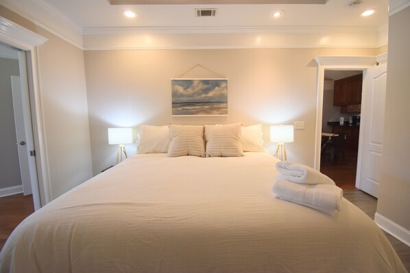A neatly made bed with pillows and a folded towel, a painting of a landscape on the wall, and a doorway leading to a kitchen area.