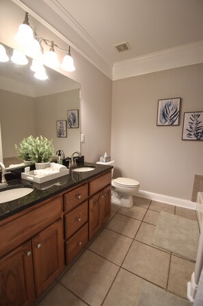 A bathroom with two sinks, a toilet, and a shower area. There are framed leaf patterns on the wall, a potted plant, and a mirror above the sinks.