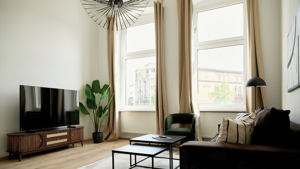 A living room with a flat-screen TV, a coffee table, a sofa, a chair, a potted plant, and a large window with curtains. - Stylish Apartment in Berlin Mitte (Berlin)