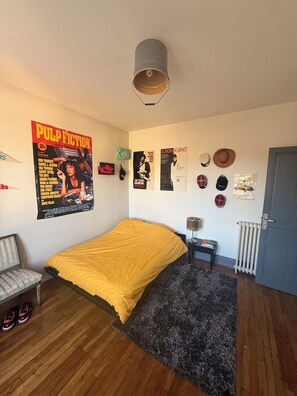 Et soveværelse med seng, radiator, dør og vægdekorationer, herunder plakater og hatte.