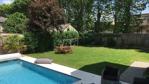 Una piscina amb una zona de terrassa, taula i cadires de fusta, i un prat amb gespa.