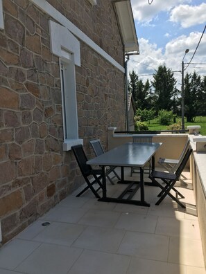 En fliseterrasse med et bord og stole, et hvidt vindue, og grøn vegetation i baggrunden.