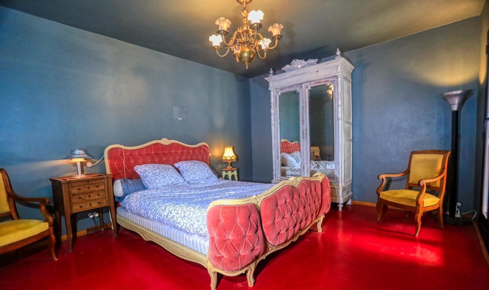 Une chambre avec un tapis rouge, un grand lit doté d’une tête de lit rouge, une armoire blanche, deux chaises et un lustre.