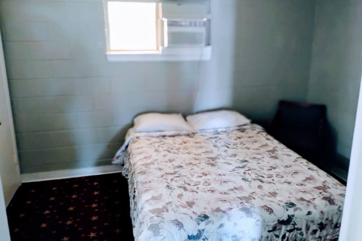 Una habitación pequeña con cama individual, colcha estampada, ventana con persianas y una alfombra oscura.