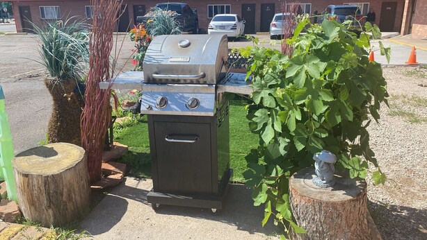 A portable gas grill with four burners, placed on a concrete surface with a tree stump nearby. - Hotel O Englewood-South Denver CO (Englewood)