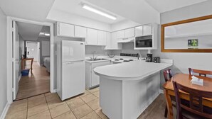 A modern kitchen with white cabinetry, a central island, and a dining area with a wooden table and chairs. - Capital O Sunset Waves Hotel Myrtle Beach Oceanfront (Myrtle Beach)
