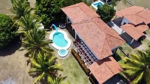 Luchtbeeld van een woonwijk met een zwembad, omringd door palmbomen, een overdekt terras en een huis met een pannendak.