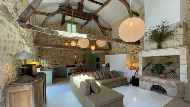 Un intérieur rustique avec des poutres apparentes, un mur en pierre, un grand canapé d’angle beige, une cheminée et des luminaires suspendus.