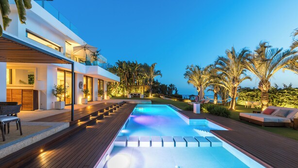 Un área de piscina moderna al aire libre con una terraza de madera, sillones de descanso y un edificio bien iluminado con grandes ventanales.