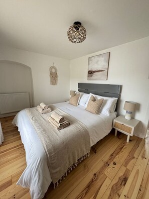 A bedroom with a bed, a bedside table, a lamp, a wall hanging, and a ceiling light fixture. - Tree Hill Cottage – Sleeps 4 | Stratton, Bude (Stratton)