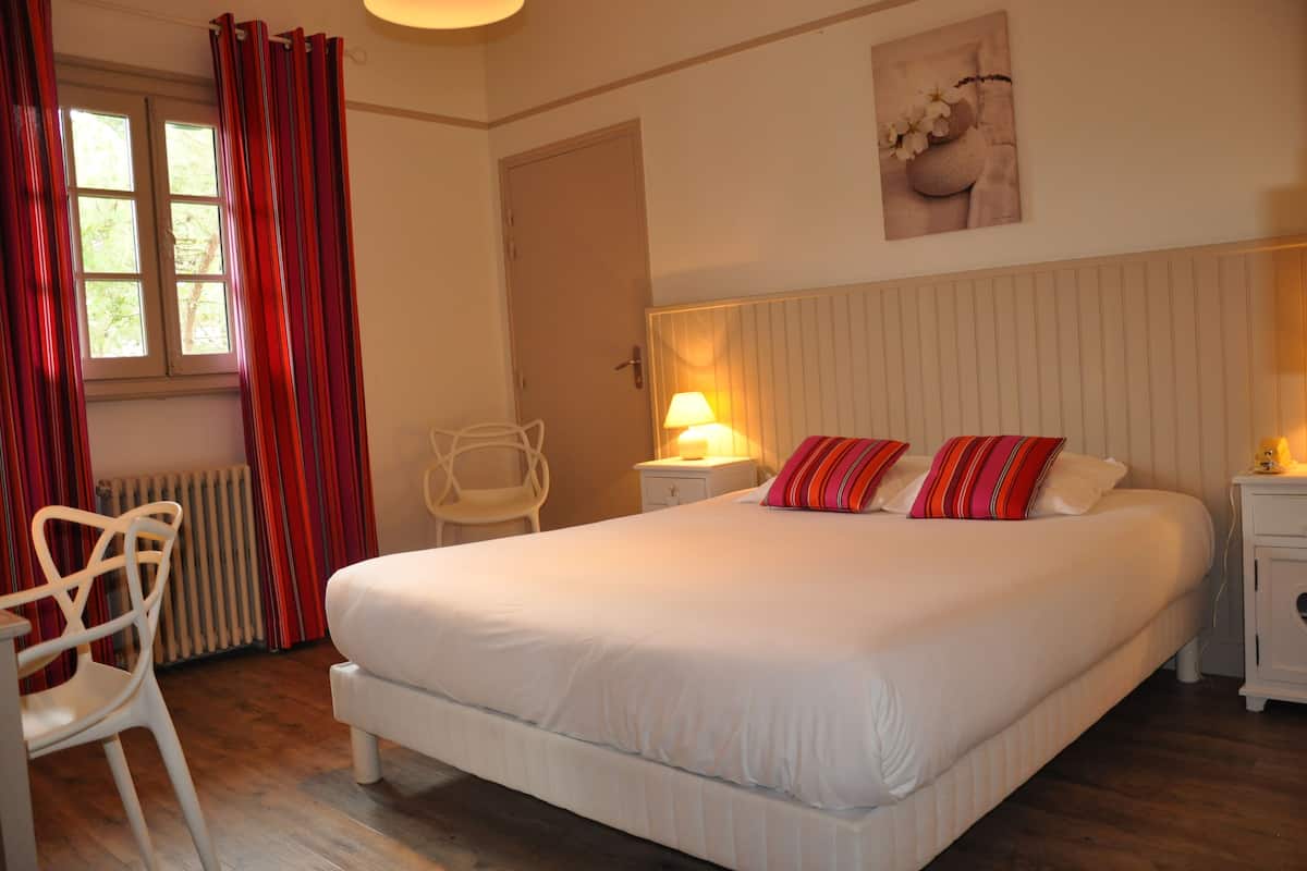 Een slaapkamer met een groot bed, een stoel, een nachtkastje en een raam met gordijnen.