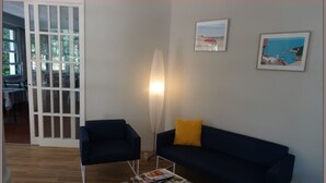 Een woonkamer met een donkerblauwe bank, een witte salontafel met tijdschriften, een geel kussen en twee fauteuilstoelen. Er is een witte deur met glaspanelen, een vloerlamp en twee ingelijste foto’s aan de muur.