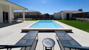 Ein rechteckiger Pool mit weißem Rand, umgeben von einer Betonterrasse. Auf der Terrasse stehen zwei Liegestühle und ein kleiner runder Tisch.
