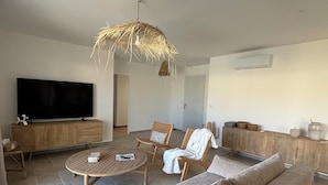 Ein Wohnzimmer mit einem großen Flachbildfernseher, einem runden Couchtisch aus Holz, zwei Korbstühlen, einem beigen Sofa und einer Hängeleuchte aus Rattan.