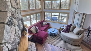 A spacious living room with a stone wall, a red sofa, a beige sofa, and a round coffee table. Large windows provide a view of the outdoors. - Family-Friendly Ski-In/Ski-Out | In-Unit Sauna (Lutsen)
