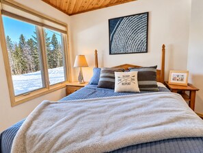 Une chambre avec un grand lit, une fenêtre donnant sur des arbres, un tableau accroché au mur et une table de chevet avec une lampe.