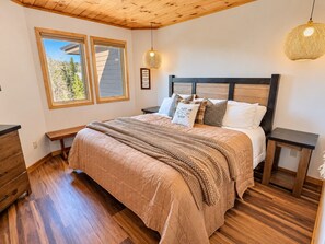 Une chambre à coucher avec un grand lit, une tête de lit en bois, deux tables de chevet, un banc et une fenêtre donnant sur des arbres.
