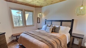 A bedroom with a wooden ceiling, a large bed with a headboard, two bedside tables, and a window with a view of trees. - Family-Friendly Ski-In/Ski-Out | In-Unit Sauna (Lutsen)