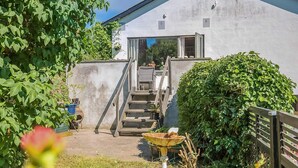 Un edificio blanco con una escalera que lleva a una puerta, rodeado de follaje y un pequeño jardín con una fuente para pájaros.