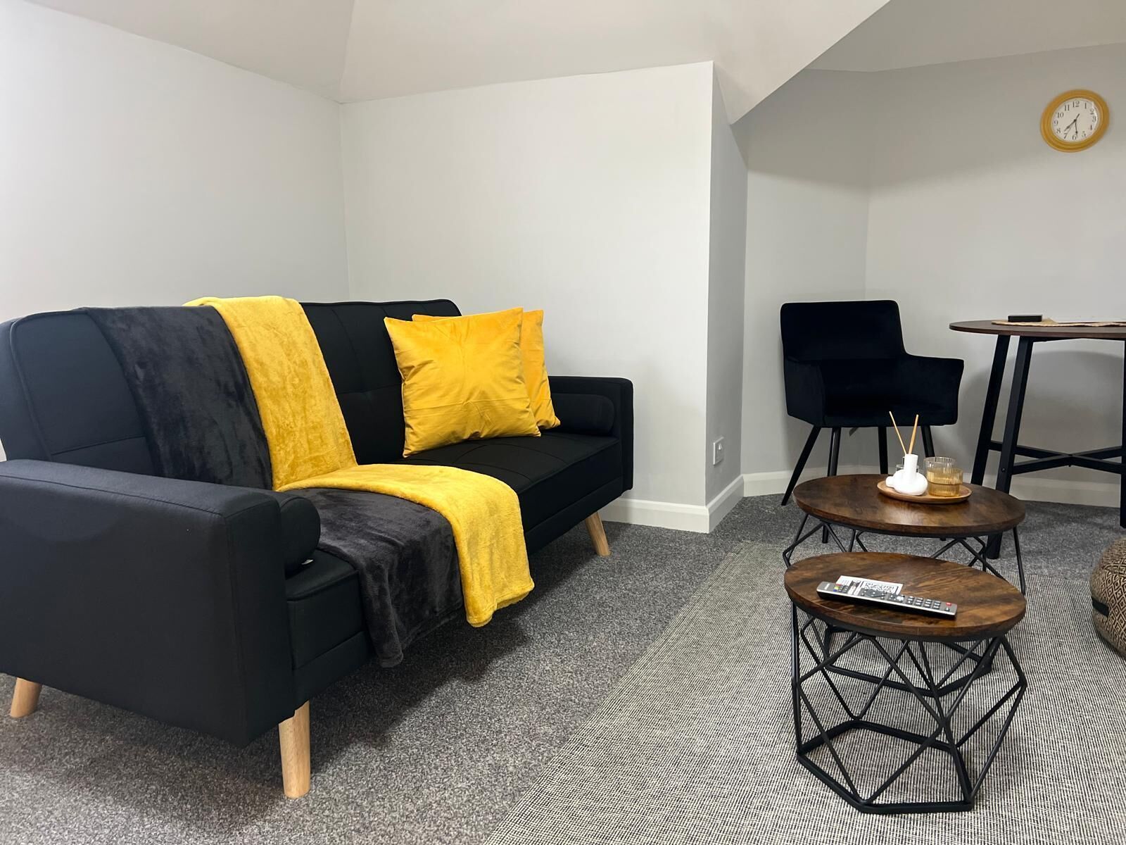 Un salon moderne avec un canapé noir, des coussins de couleur jaune, une table basse ronde et une petite table d’appoint.