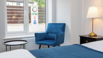 A hotel room with a blue armchair, a bedside table with a lamp, a bed, and a window with a view of a fence and a building.