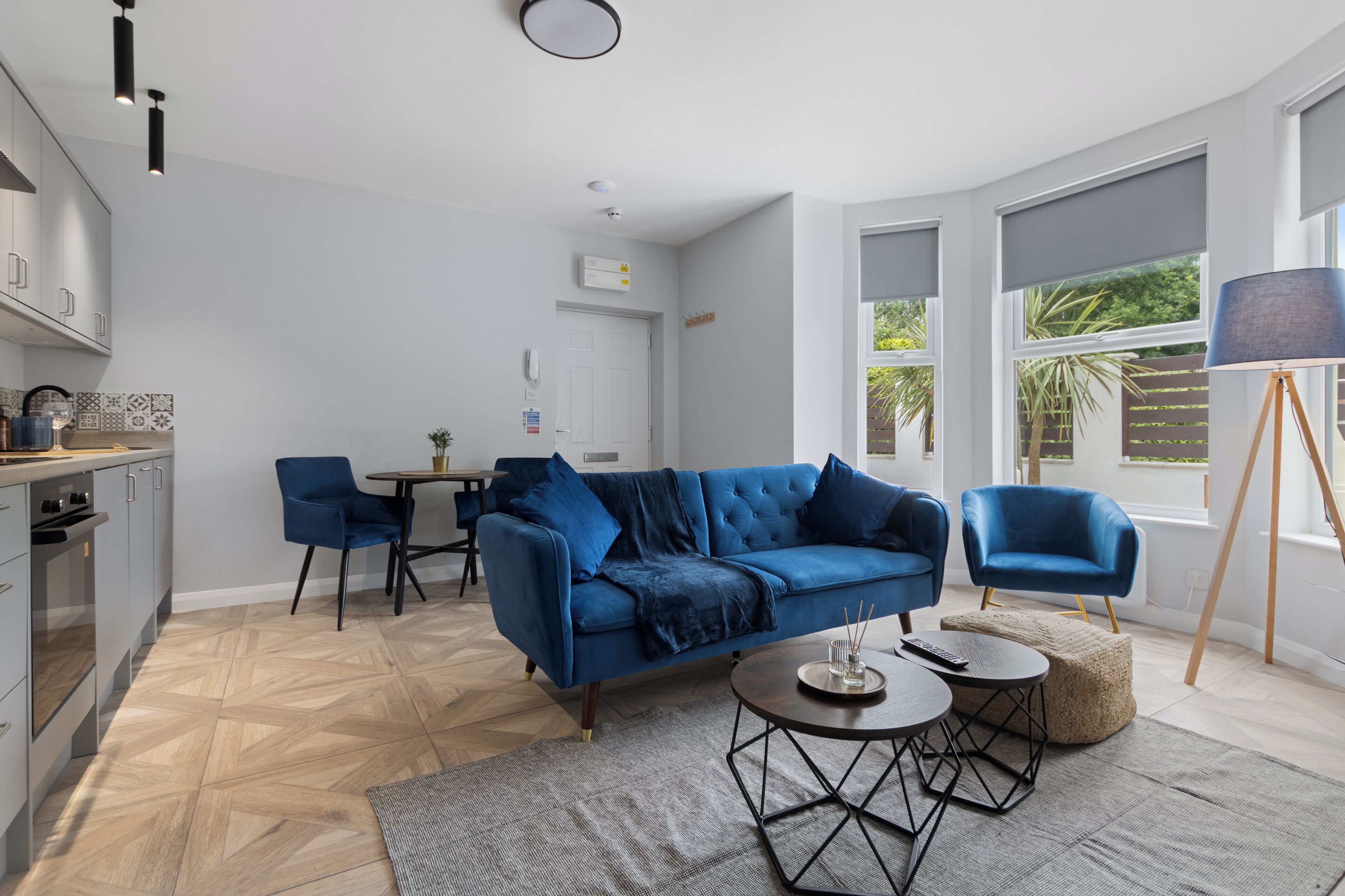 A modern living room with a blue sofa, a dining table, and a kitchen area with grey cabinets.