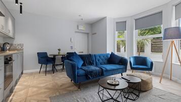 A modern living room with a blue sofa, a dining table, and a kitchen area with grey cabinets.