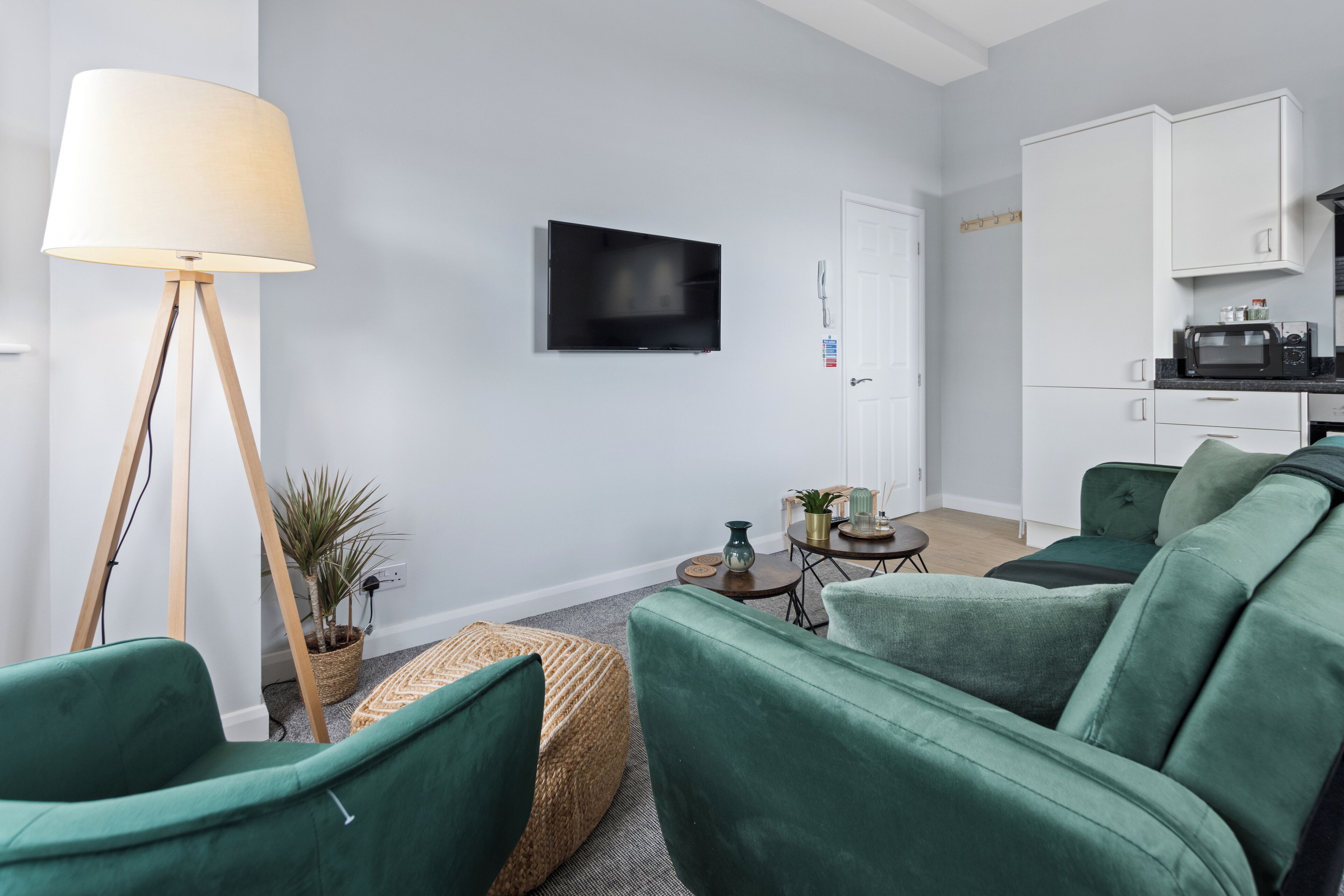 Ein modernes Wohnzimmer mit Fernseher, grünen Sofas, einer Stehlampe und einer kleinen Küchenzeile.