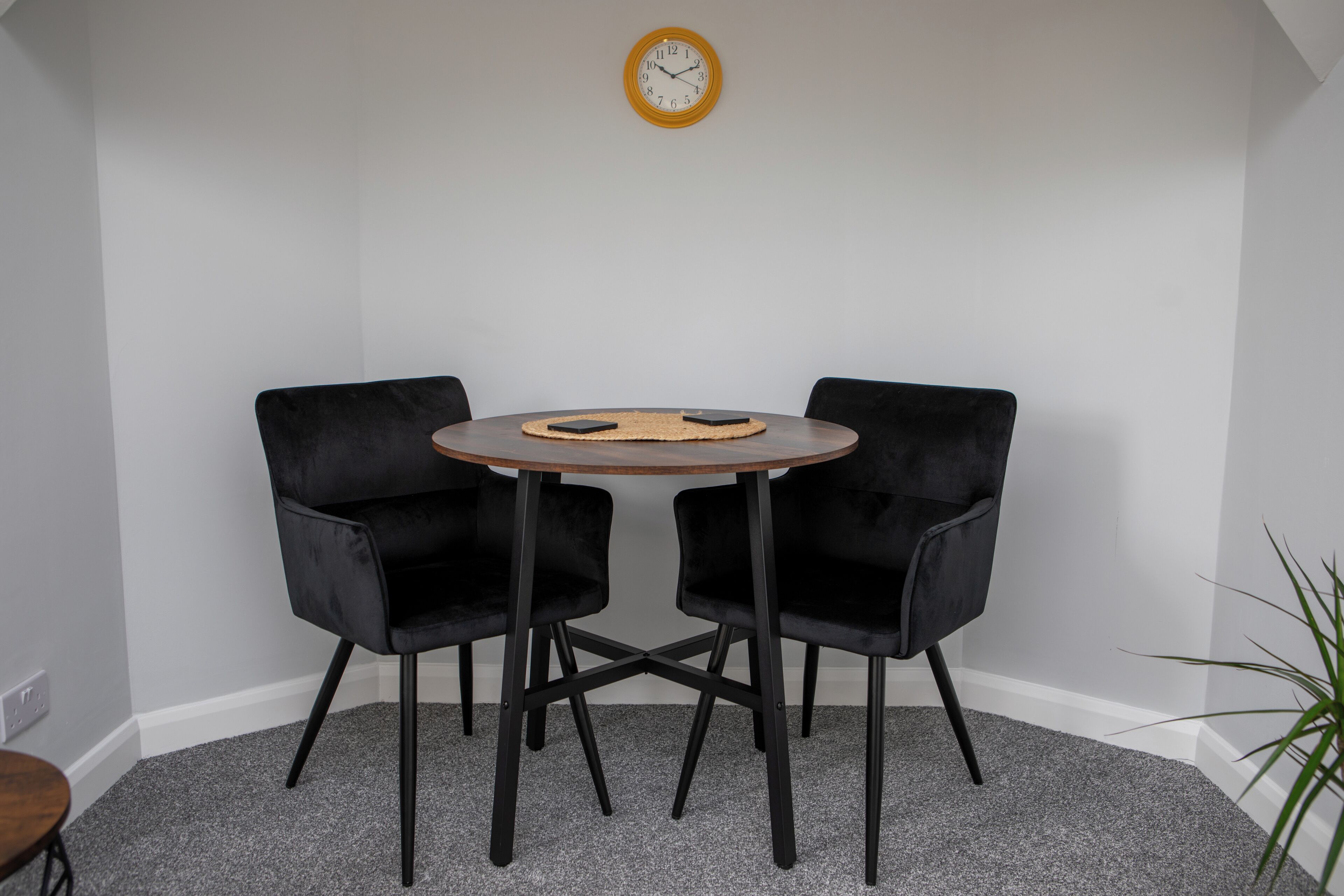 Una habitación con una mesa redonda, dos sillas negras, un reloj en la pared y una planta en la esquina.
