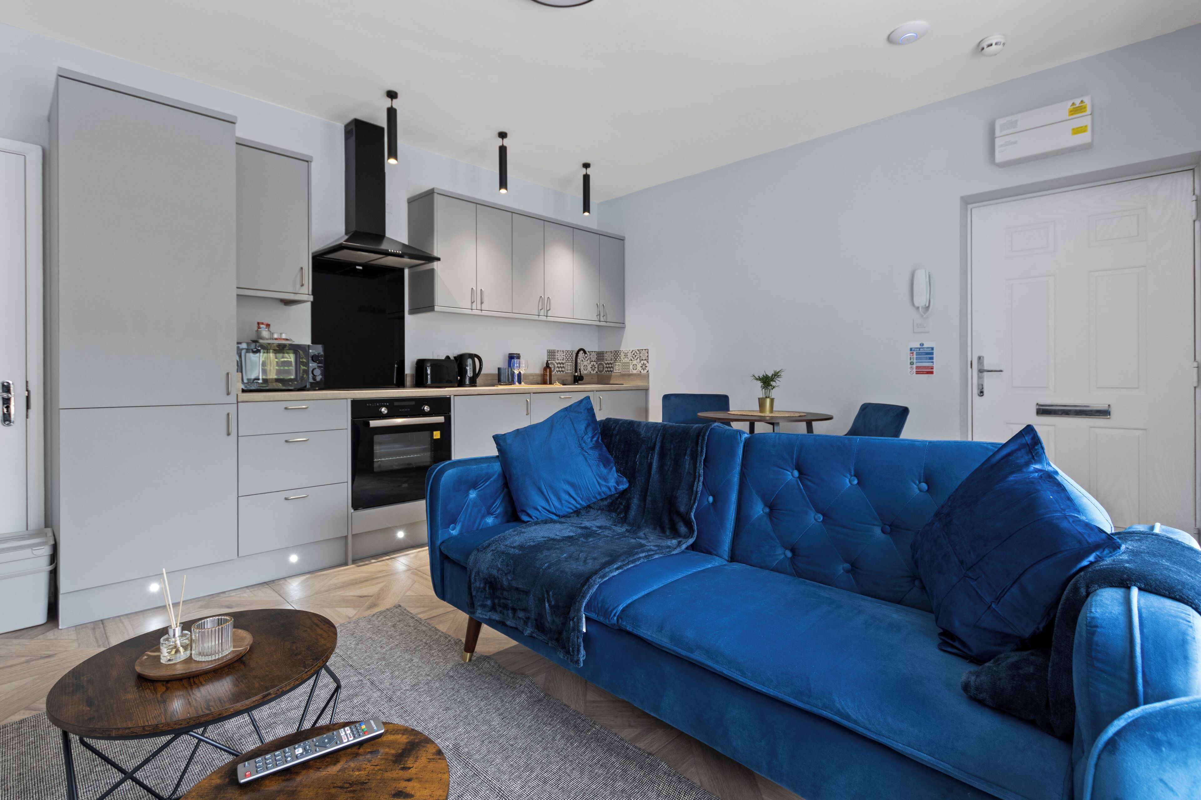 Una sala de estar moderna con un sofá azul, una mesa de centro y una cocina con armarios grises y una campana extractora negra.