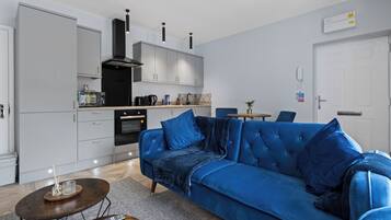 Una sala de estar moderna con un sofá azul, una mesa de centro y una cocina con armarios grises y una campana extractora negra.
