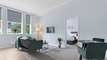 Un salon moderne avec un canapé, une table à manger et des chaises, un téléviseur fixé au mur, et un lit dans la pièce adjacente.