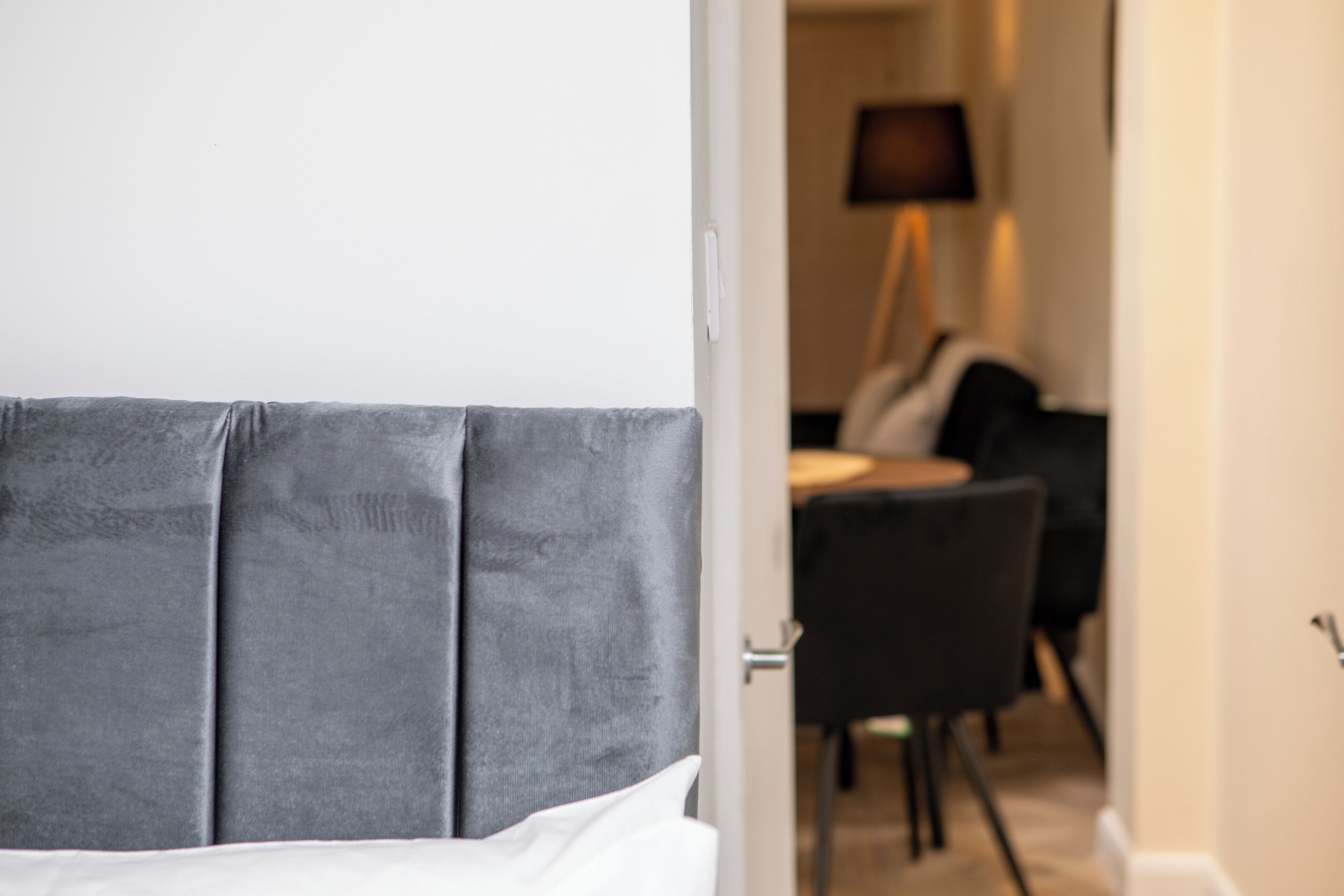 Habitación de hotel con cabecera tapizada en gris, almohada blanca y puerta que da a un comedor con mesa y sillas.