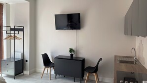 Een moderne, minimalistisch ingerichte kamer met een flatscreen televisie aan de muur, een klein bureau met stoel, een potplant en een geïntegreerd keukenhoekje met spoelbak en kookplaat.