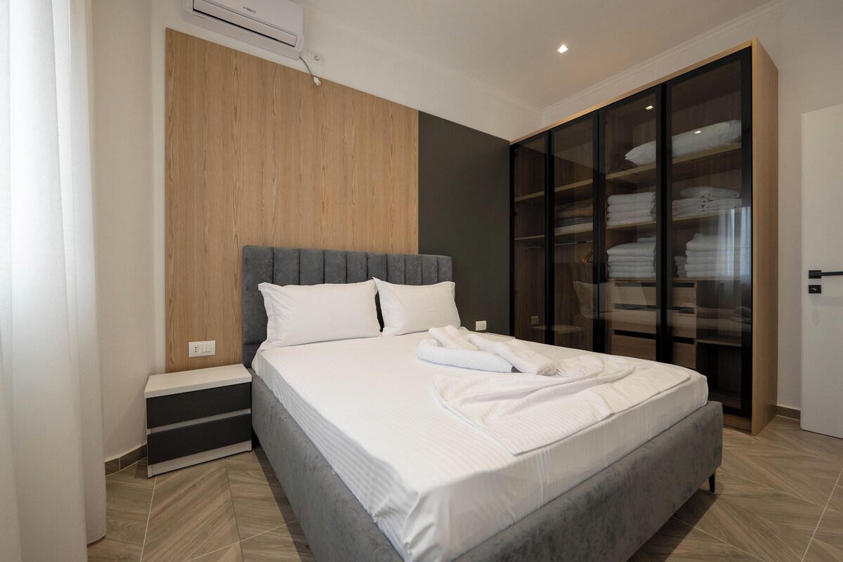 A modern bedroom with a large bed, a wooden accent wall, a glass wardrobe, and a nightstand.