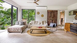 A modern living room with a bed, sofa, and coffee table, large windows, and a view of greenery.