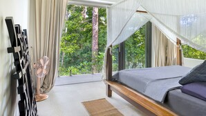 A bedroom with a bed, a standing fan, and a view of greenery outside.