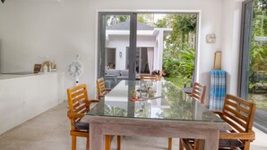 A modern dining area with a glass table and wooden chairs, a fan, and a view of a house and greenery outside.