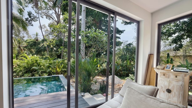 A modern living room with a large sliding glass door leading to a pool area surrounded by lush greenery.