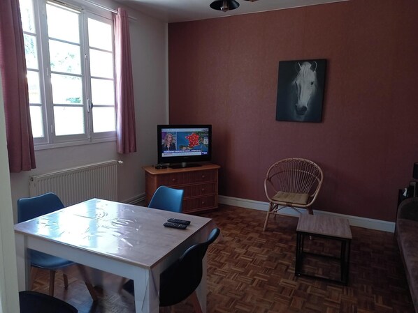 A room with a wooden floor, a table with chairs, a television, and a framed picture on the wall.