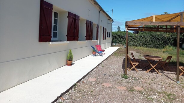 Un bâtiment blanc avec des volets rouges, un petit jardin agrémenté d’un pot de fleurs et des sièges extérieurs placés sous une pergola en bois.