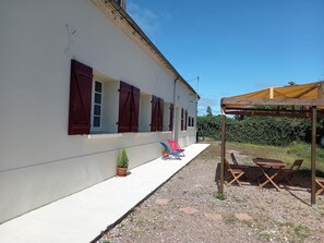 A white building with red shutters, a small garden with a potted plant, and outdoor seating under a wooden pergola.